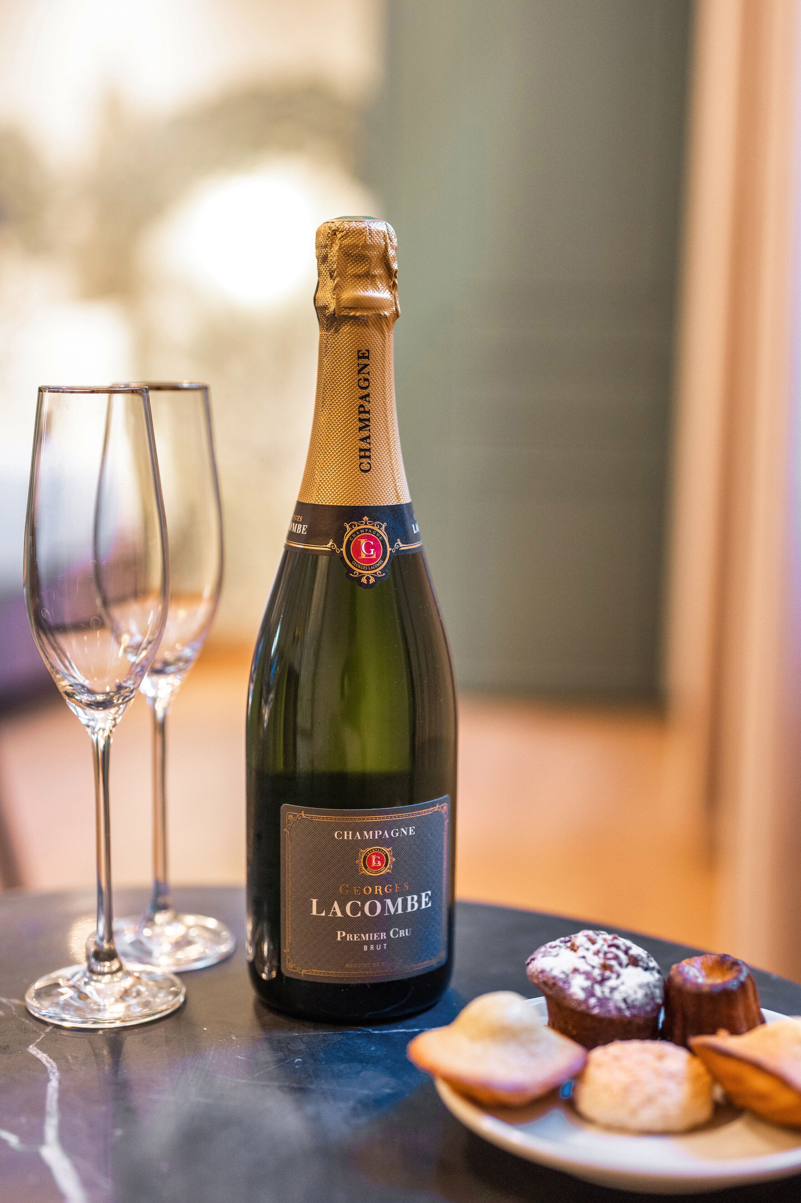 Una botella de champán George La Côme Premier Cru Brut, dos copas de vino vacías y un plato con pasteles sobre una mesa.