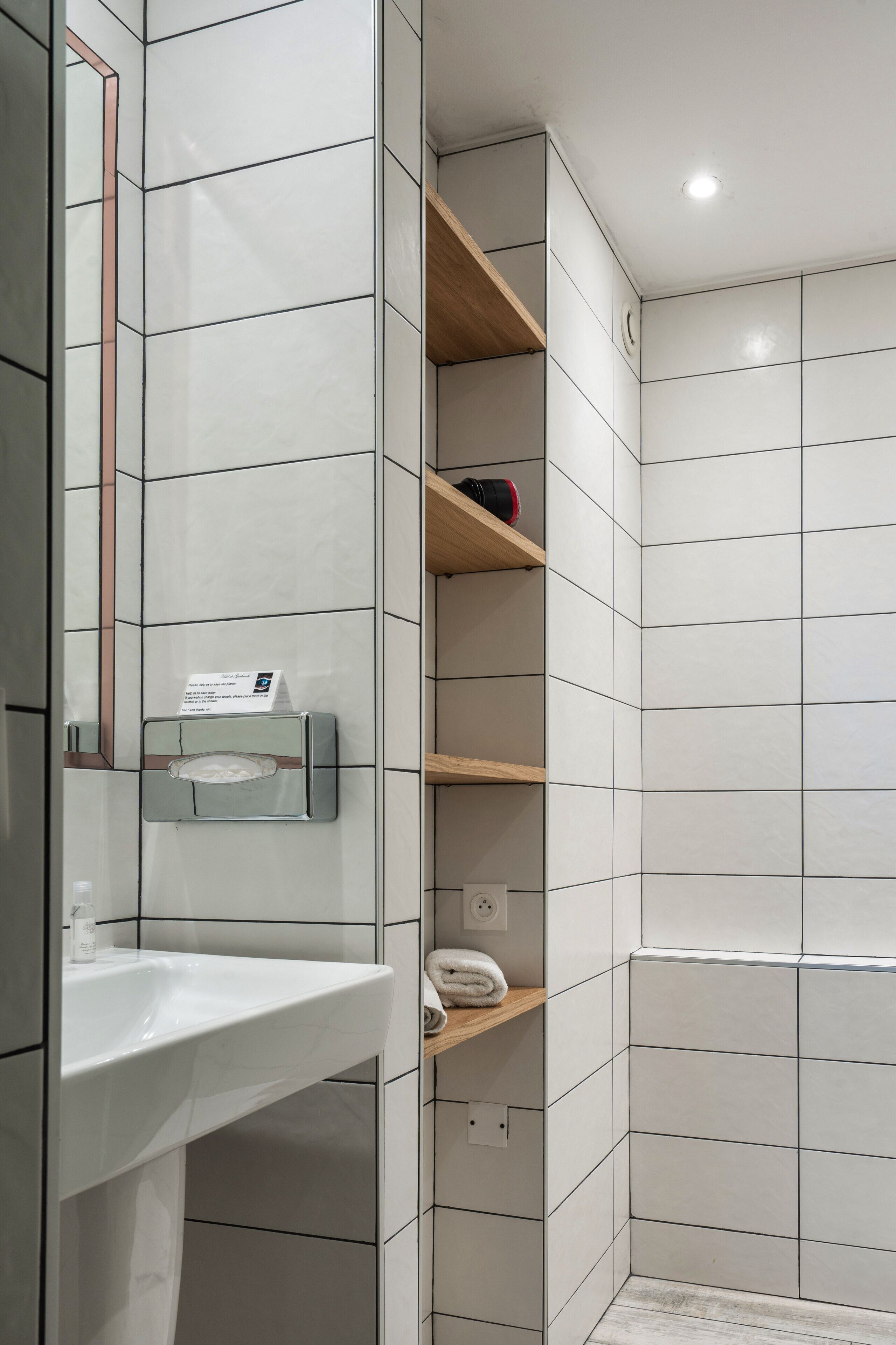 Une salle de bain avec des murs recouverts de carreaux blancs, un lavabo blanc et des étagères en bois.