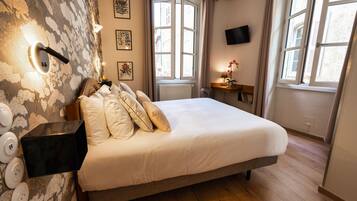 A bedroom with a bed, a wall-mounted light, a radiator, a window, a TV, and a small table with a lamp and flowers.