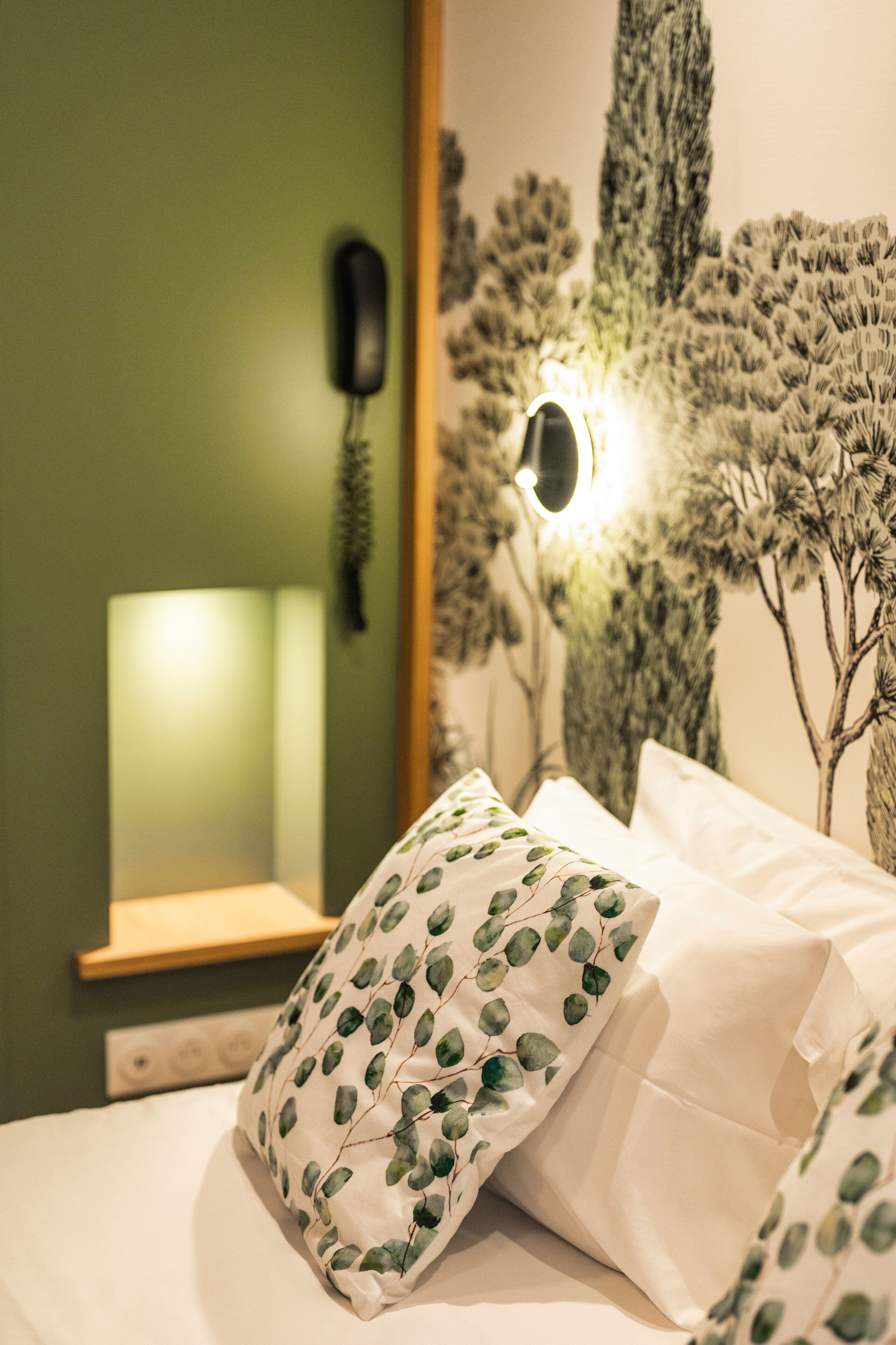 A bedroom with a green wall, a bedside lamp, a wall-mounted phone, and a decorative tree mural.