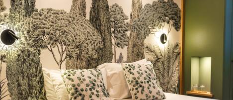 Una cama con ropa de cama blanca y almohadas estampadas, una pared con un mural de un árbol y una pequeña estantería con vasos.