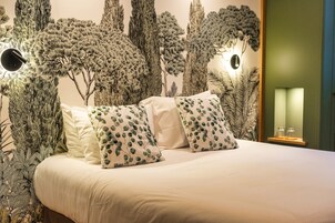 Una cama con ropa de cama blanca y almohadas estampadas, una pared con un mural de un árbol y una pequeña estantería con vasos.