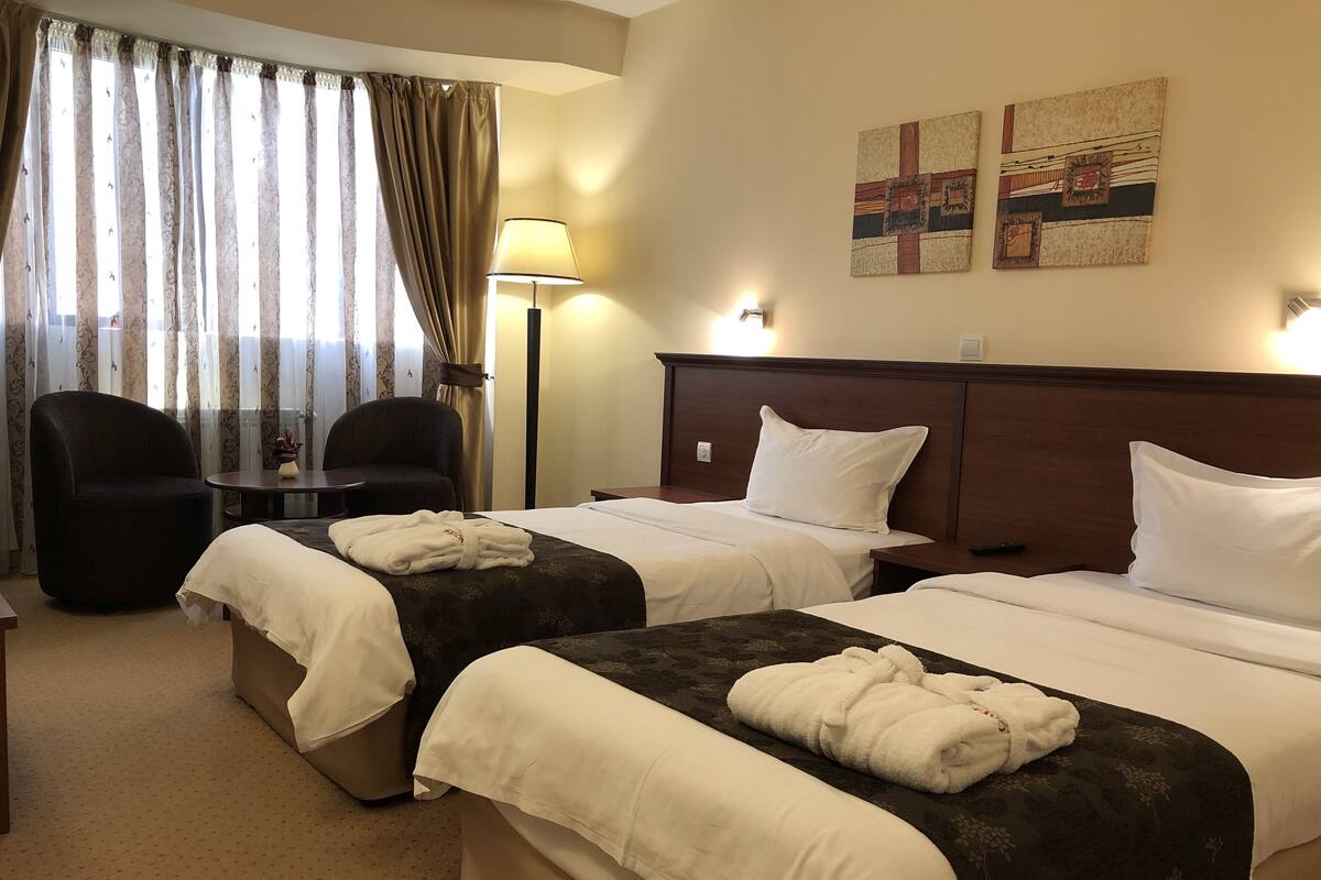 A hotel room with two beds, a desk with a chair, a lamp, and a window with curtains.
