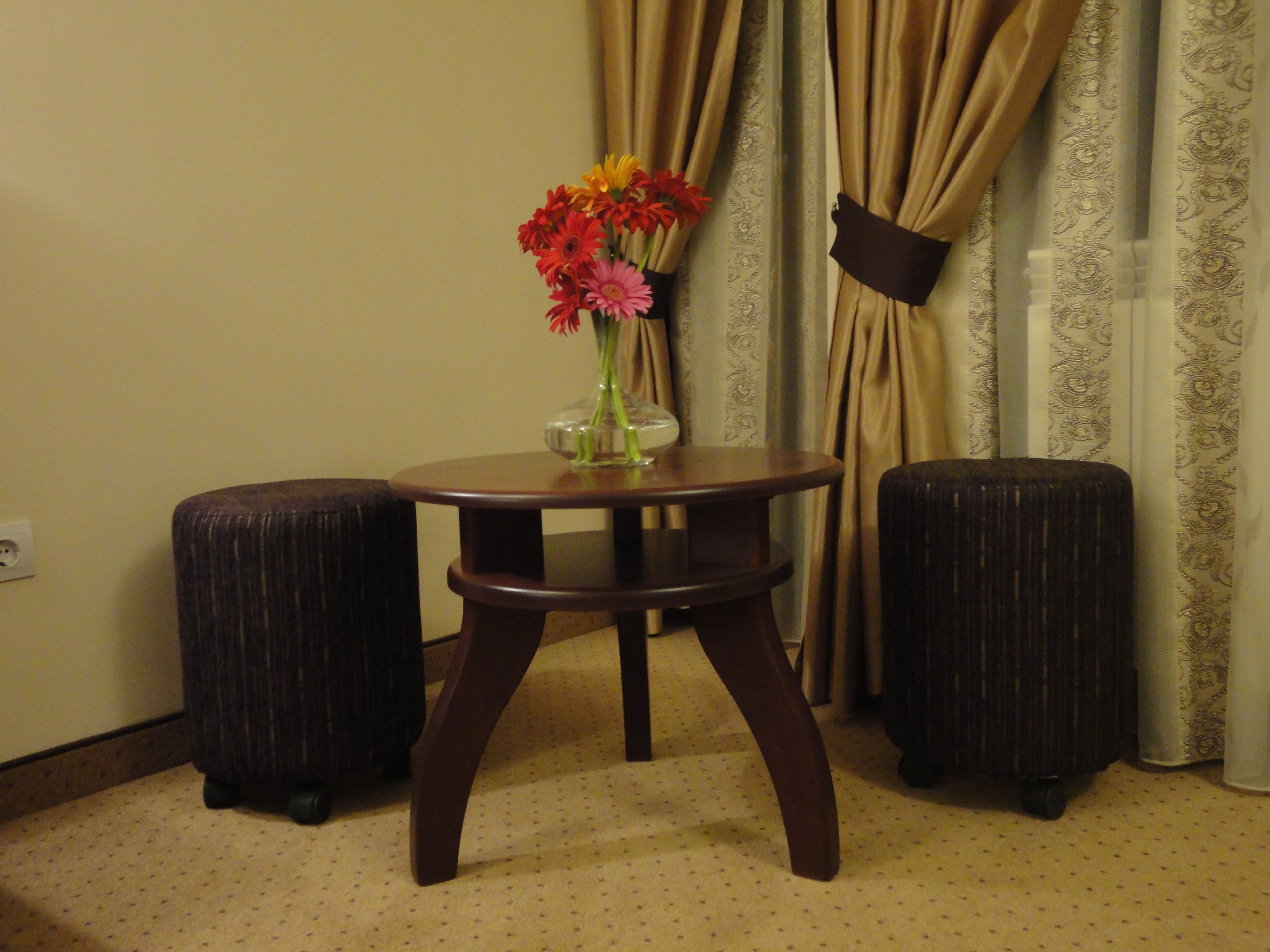 Une petite table ronde surmontée d’un vase de fleurs, deux poufs et un rideau dans une pièce dont le mur et le tapis sont de couleur unie.