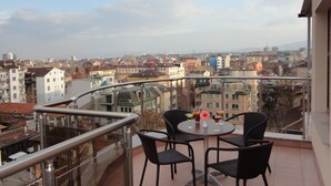 A balcony with a table and chairs, overlooking a cityscape with various buildings and a distant mountain range. - Hotel Favorit (Sofia)