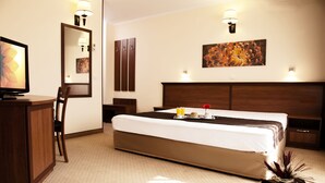 A hotel room with a bed, bedside table, lamp, artwork, and a desk with a chair. - Hotel Favorit (Sofia)