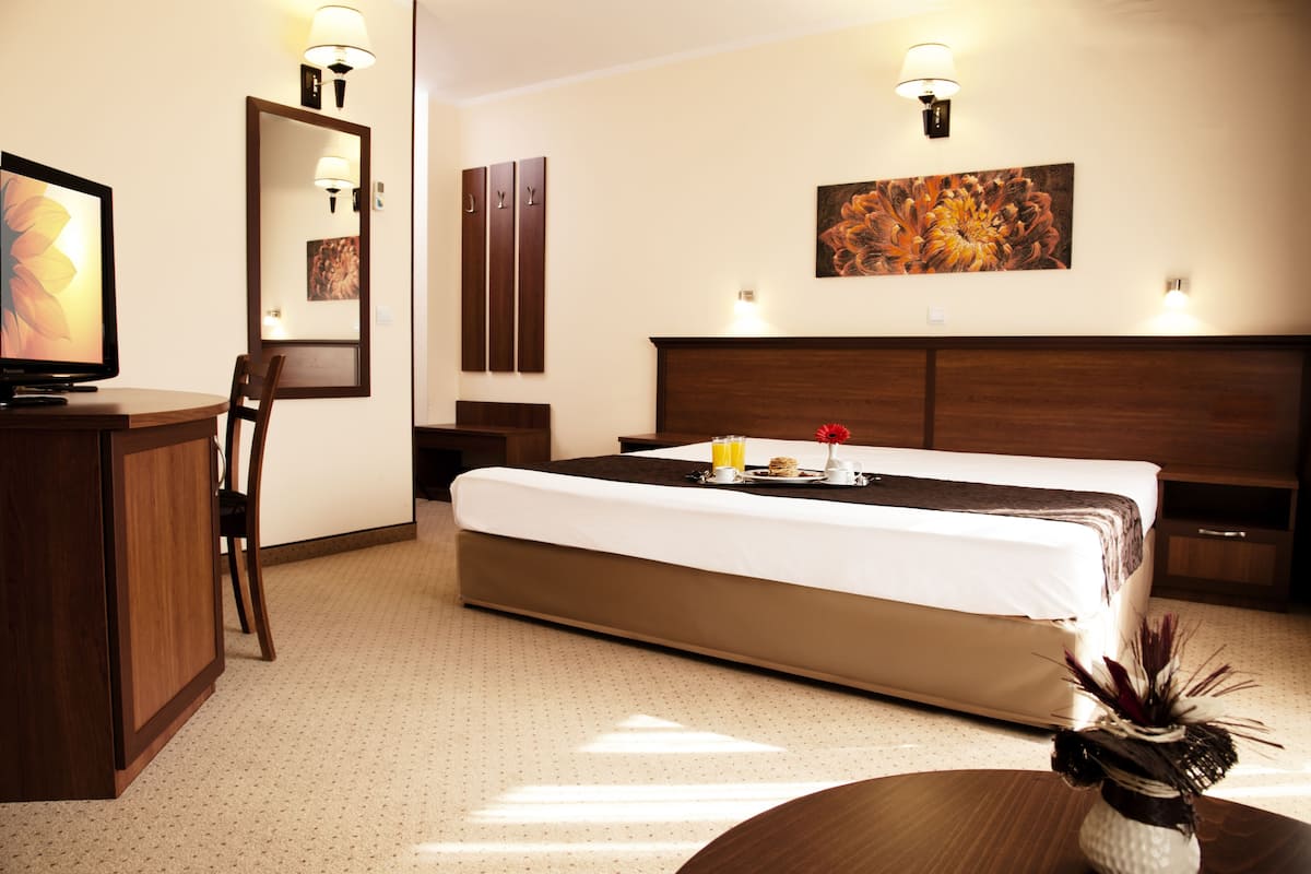 A hotel room with a bed, bedside table, lamp, artwork, and a desk with a chair.