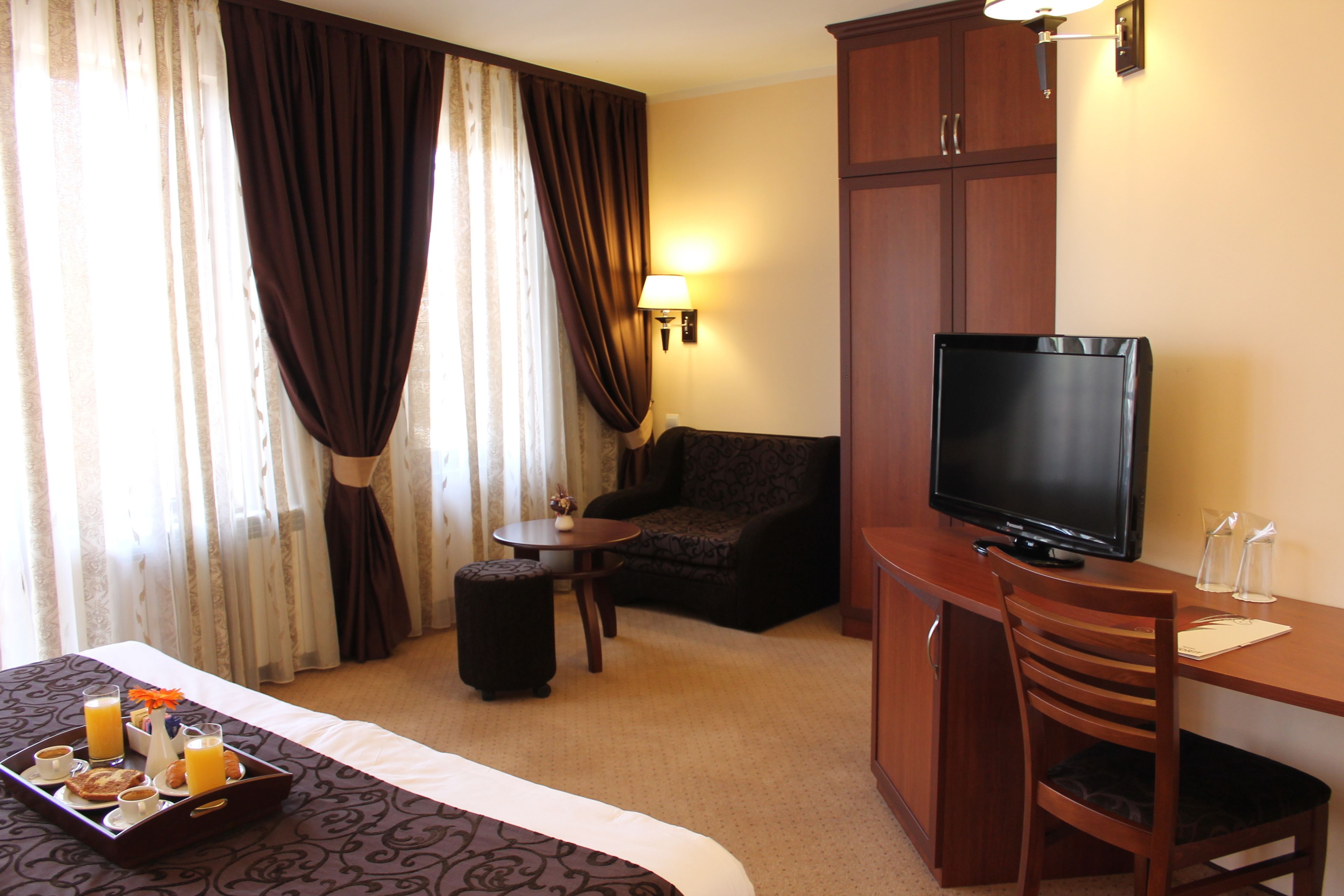 A hotel room with a bed, a TV, a desk, a chair, a small table, and a tray with breakfast items.