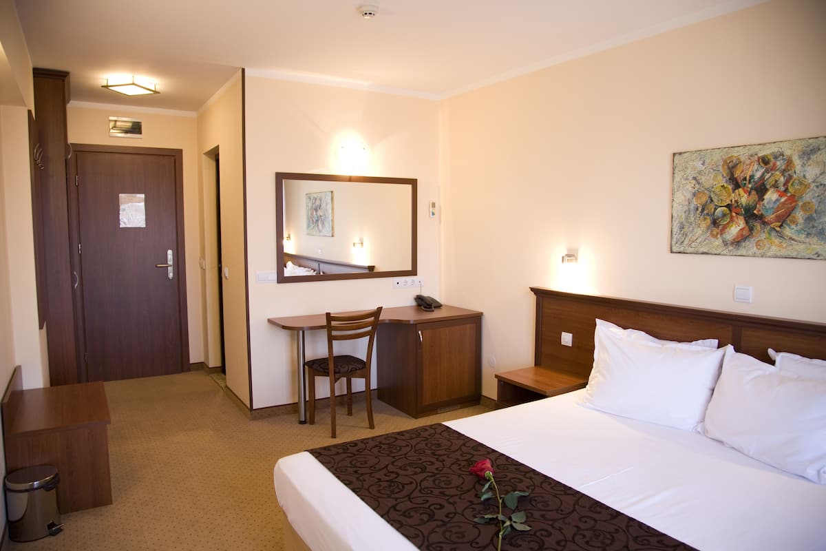 A hotel room with a bed, a desk, a chair, a small cabinet, a trash bin, and a painting on the wall.