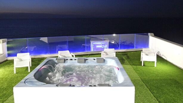 A hot tub with a glass enclosure, surrounded by white chairs, set on artificial grass with a view of the sea. - Hotel Residence La Baia (Castelsardo)