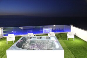A hot tub with a glass enclosure, surrounded by white chairs, set on artificial grass with a view of the sea. - Hotel Residence La Baia (Castelsardo)