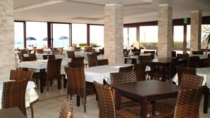 A restaurant with multiple tables and wicker chairs, large windows, and a stone pillar. - Hotel Residence La Baia (Castelsardo)
