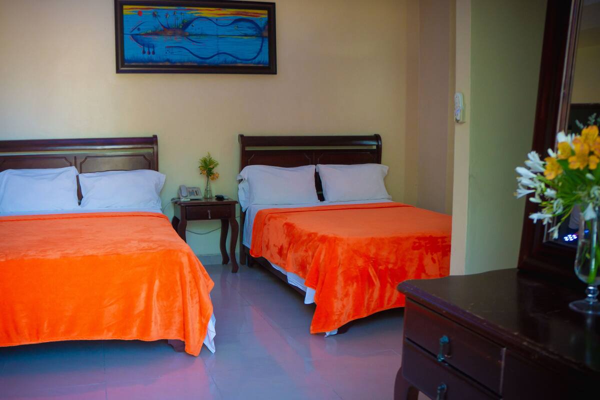A hotel room with two beds, each with an orange bedspread, a wooden headboard, and white pillows. There is a small table with a phone and a vase with flowers. A painting hangs on the wall above the beds.
