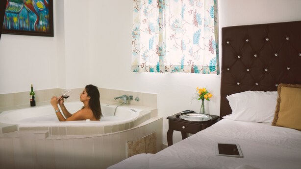 A room with a bed, a bedside table with a tablet, a window with floral curtains, a painting on the wall, and a bathtub with a bottle of wine.