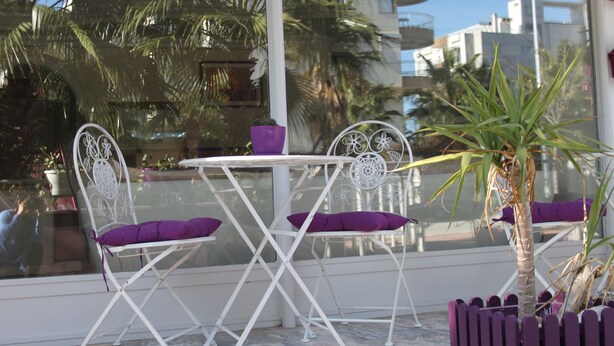 A patio area with a white metal table and chairs, each with a purple cushion. There is a potted plant in the corner and a view of palm trees and buildings through a glass window. - Villa Granada Hotel (Konyaalti)