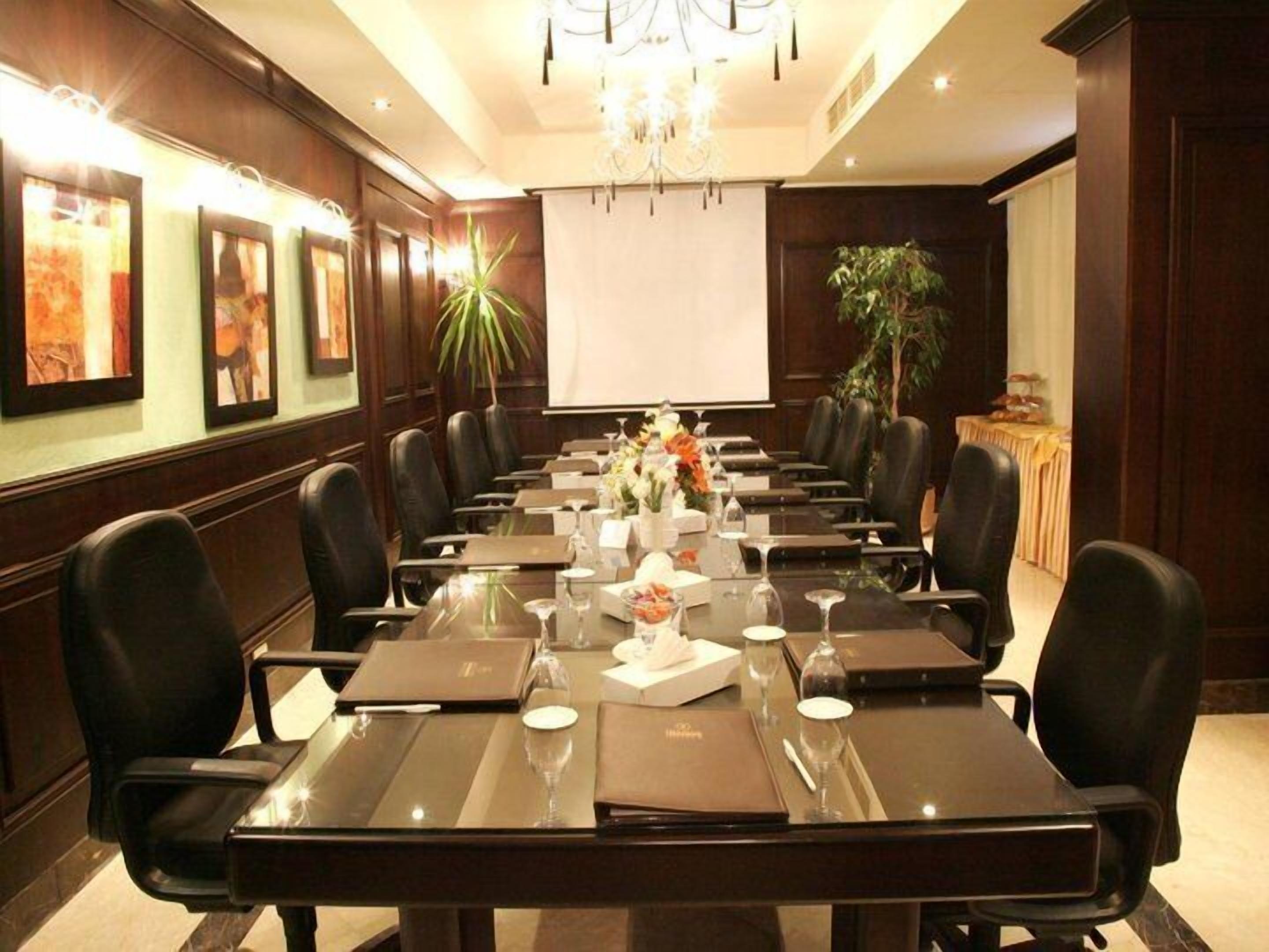 A conference room with a long table, black leather chairs, a projector screen, and a plant.