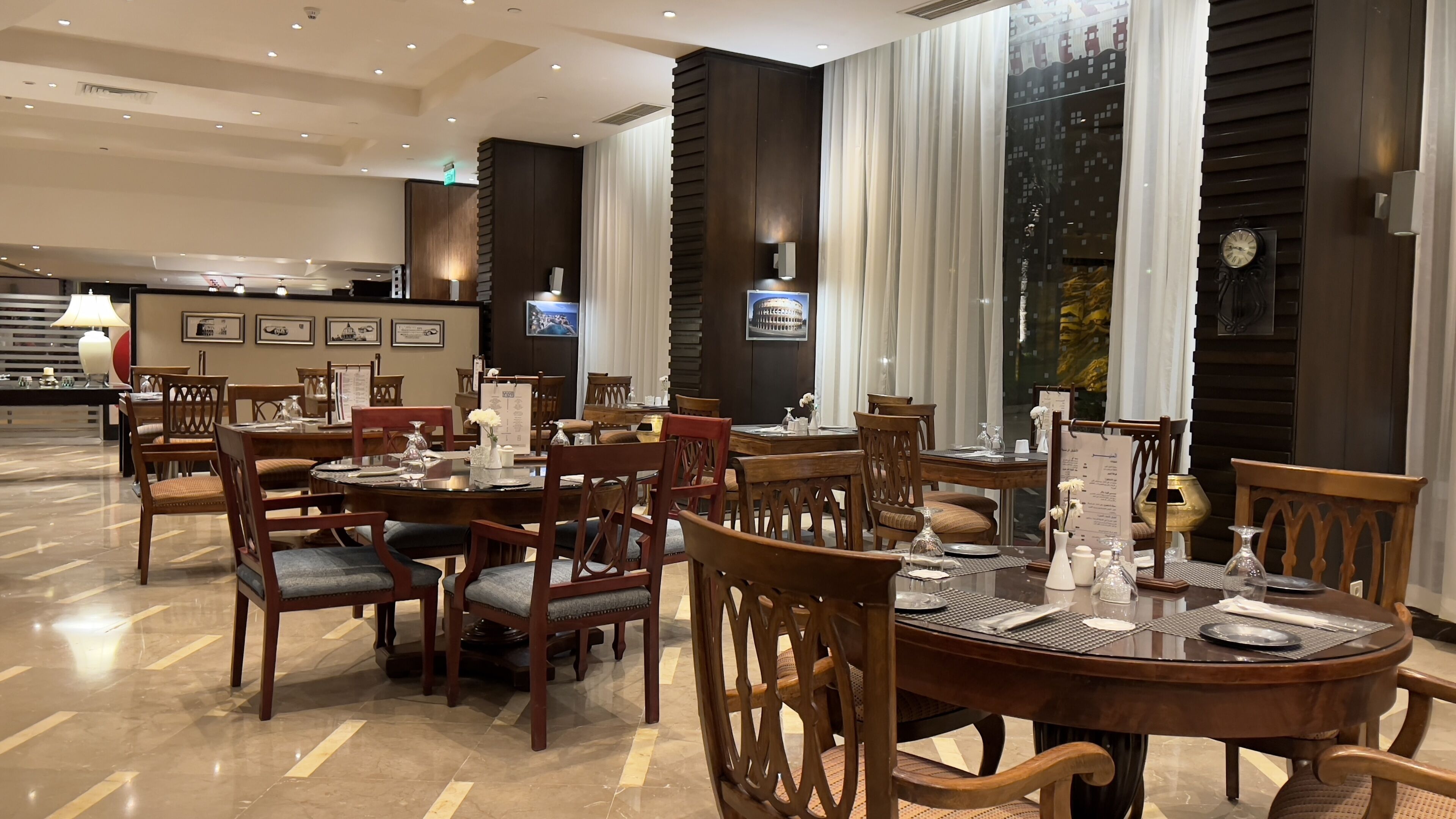 Un comedor de hotel con mesas redondas, sillas de madera y un piso de mármol pulido.
