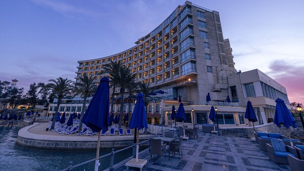 Ein Hotel mit geschwungener Fassade, mehreren Balkonen und einem Poolbereich mit Liegestühlen und Sonnenschirmen.