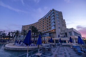 Un hotel con fachada curva, varios balcones y una zona de piscina con sillones y sombrillas.