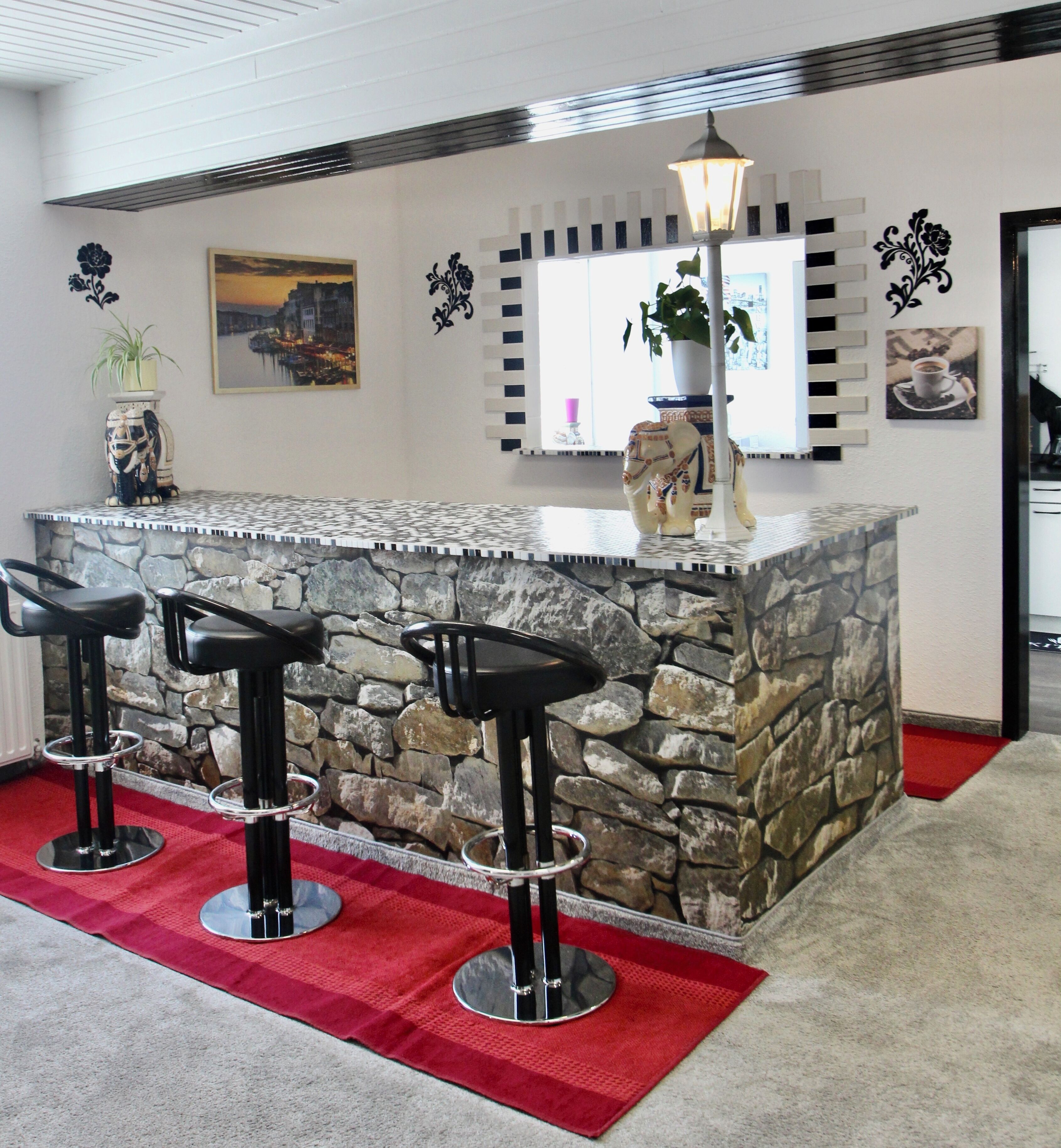 A bar area with a stone counter, three black bar stools, a red rug, and decorative wall art.