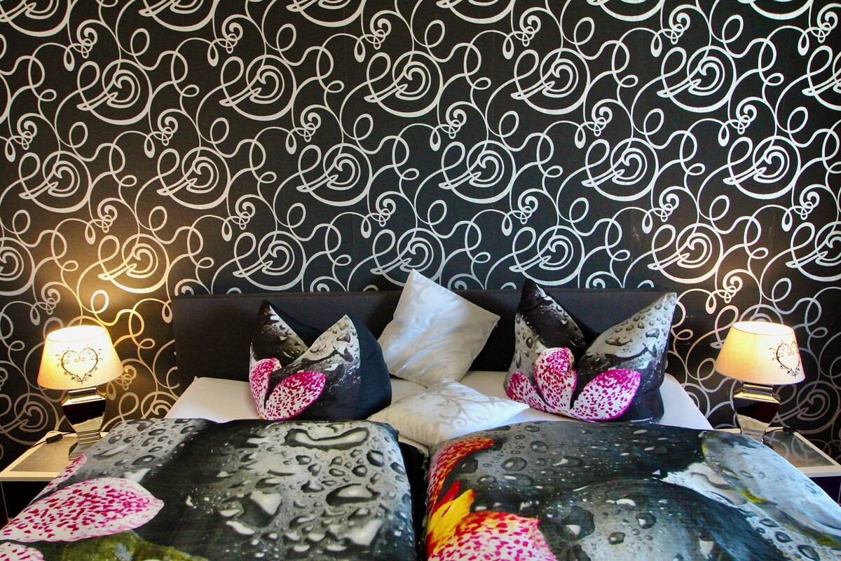 Une chambre d’hôtel avec un papier peint à motifs noir et blanc, un lit recouvert d’une couette aux motifs floraux et aquatiques, et deux lampes de chevet.