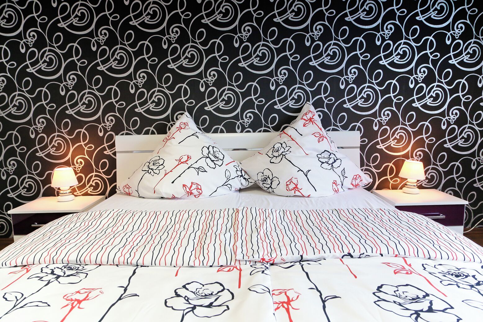 A neatly made bed with floral-patterned bedding and pillows, a bedside table with a lamp, and a wall with a black and white swirl pattern.