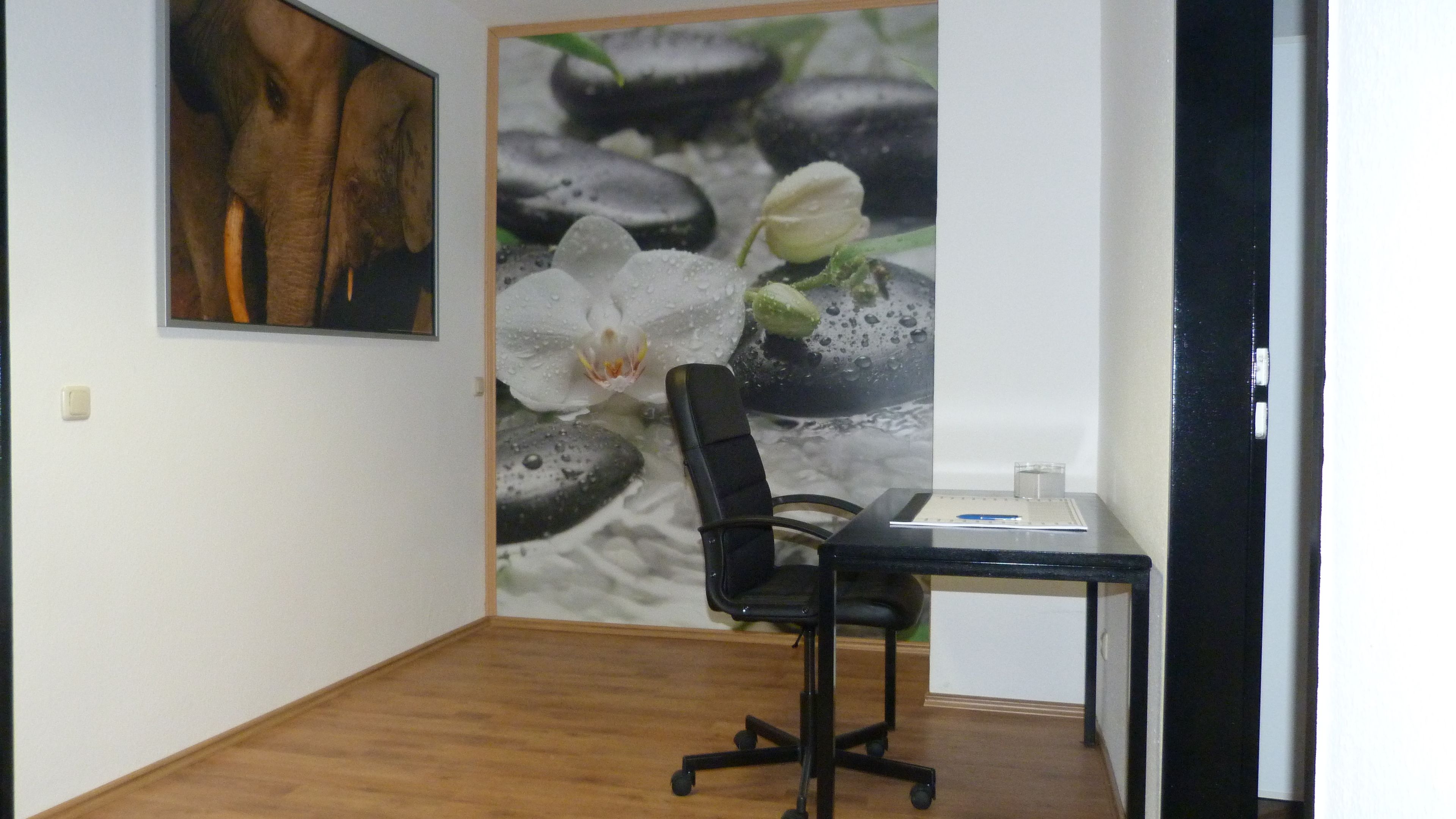 Ein Zimmer mit Holzboden, einem schwarzen Bürostuhl, einem Schreibtisch mit Computer und einer kleinen Pflanze. An der Wand hängt ein gerahmtes Bild und zur Rechten befindet sich eine Tür.