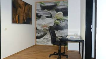 Ein Zimmer mit Holzboden, einem schwarzen Bürostuhl, einem Schreibtisch mit Computer und einer kleinen Pflanze. An der Wand hängt ein gerahmtes Bild und zur Rechten befindet sich eine Tür.