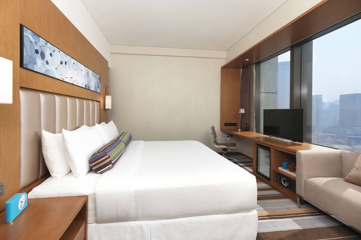 Ein modernes Hotelzimmer mit einem großen Bett, einem Fernseher, einer Sitzecke und Blick auf die Stadt.