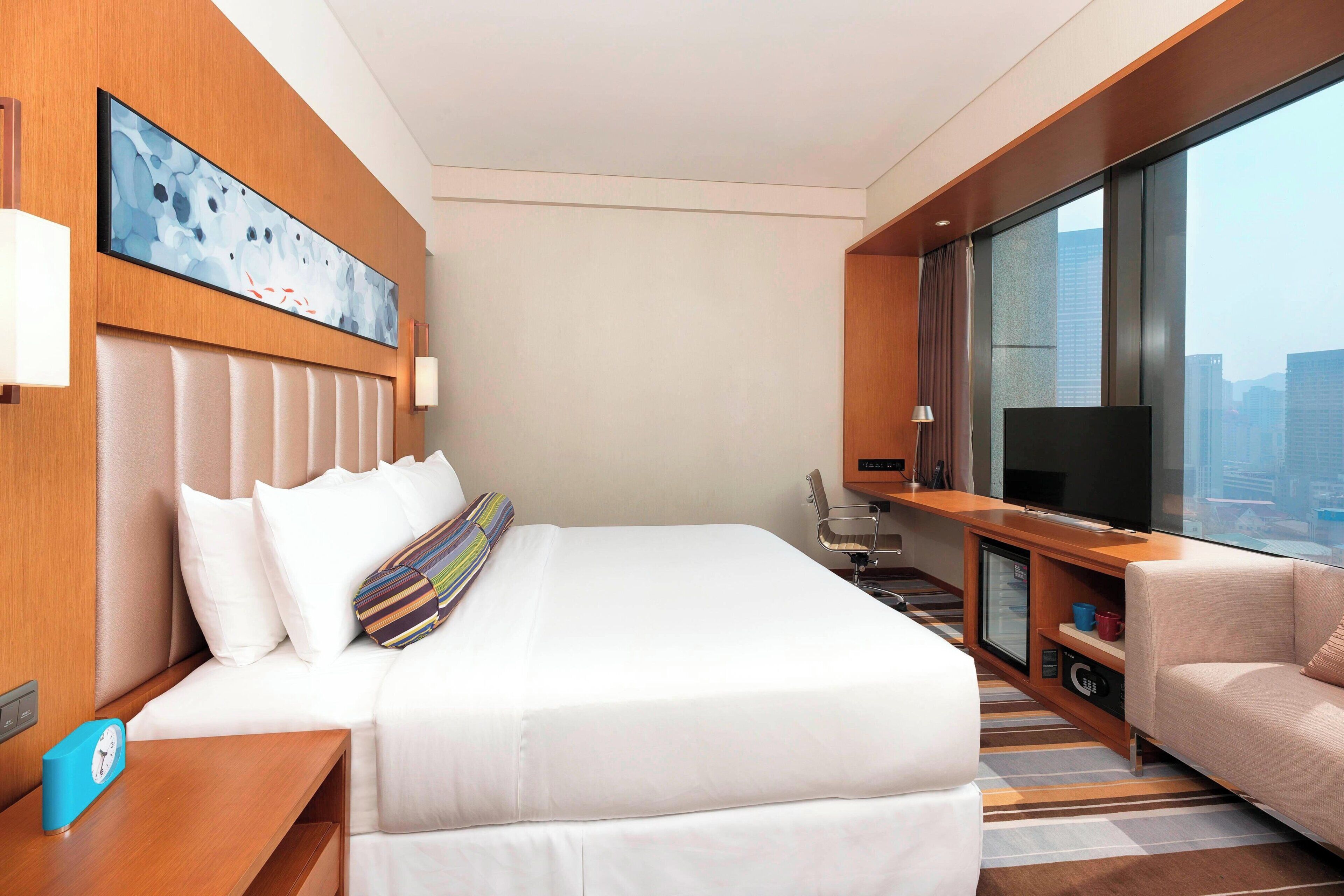 A hotel room with a large bed, a desk with a television, a sofa, and a view of the city.