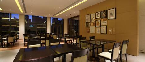 A modern dining area with tables and chairs, large windows, and framed artwork on the walls.