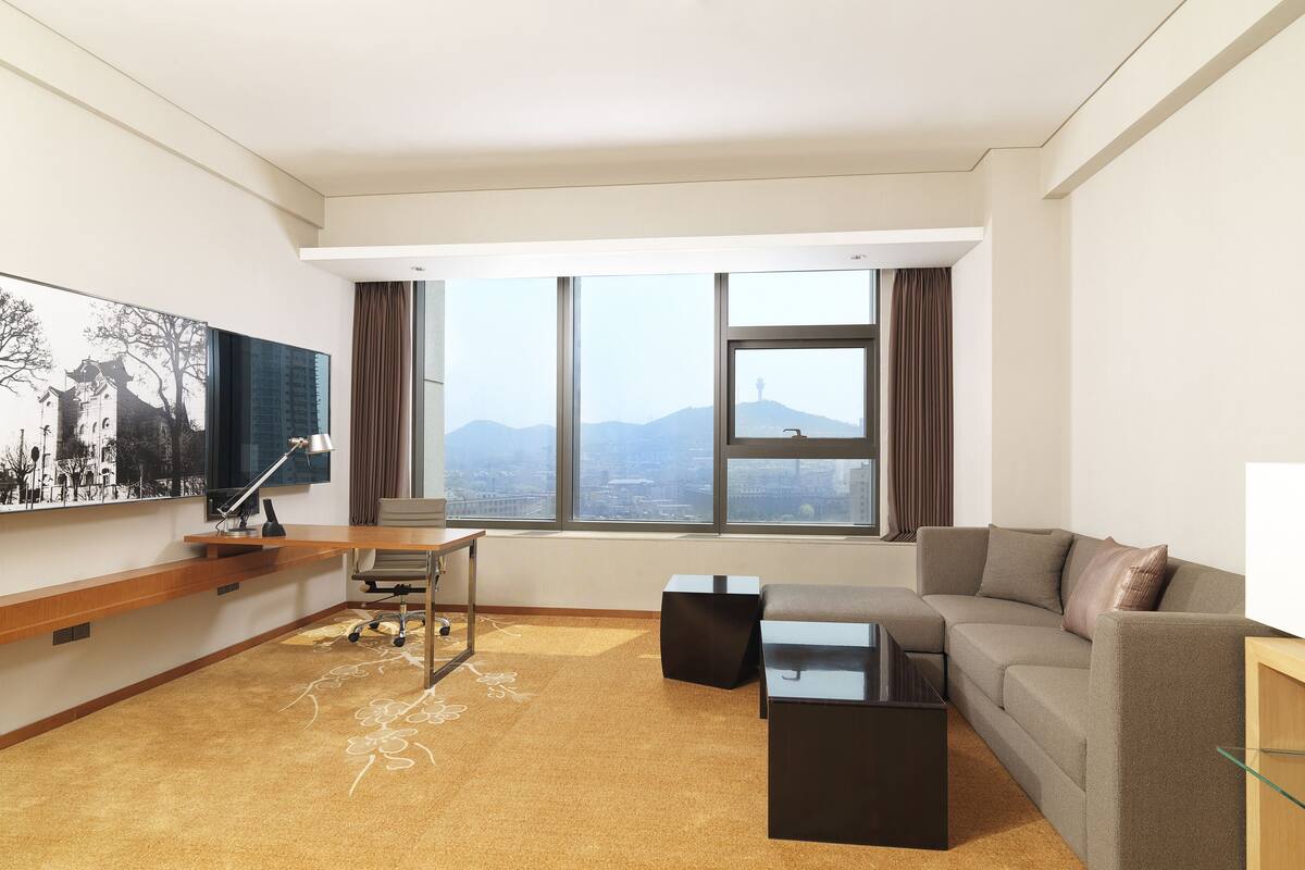 Ein modernes Hotelzimmer mit großem Fenster und Blick auf die Stadt, einem gemütlichen Sofa, einem Schreibtisch mit Lampe und einem an der Wand befestigten Flachbildfernseher.