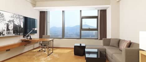 Quarto de hotel moderno com uma janela grande que oferece vista para a cidade, um sofá confortável, uma mesa com abajur e uma televisão de tela plana na parede.