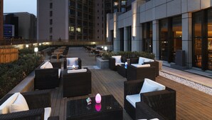 An outdoor seating area with wicker furniture and a wooden deck, surrounded by high-rise buildings. - Aloft Dalian (Dalian)