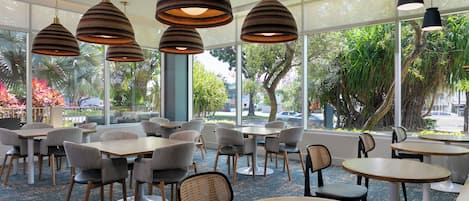 A modern indoor dining area with multiple tables and chairs, large windows with a view of greenery and a street, and pendant lighting.