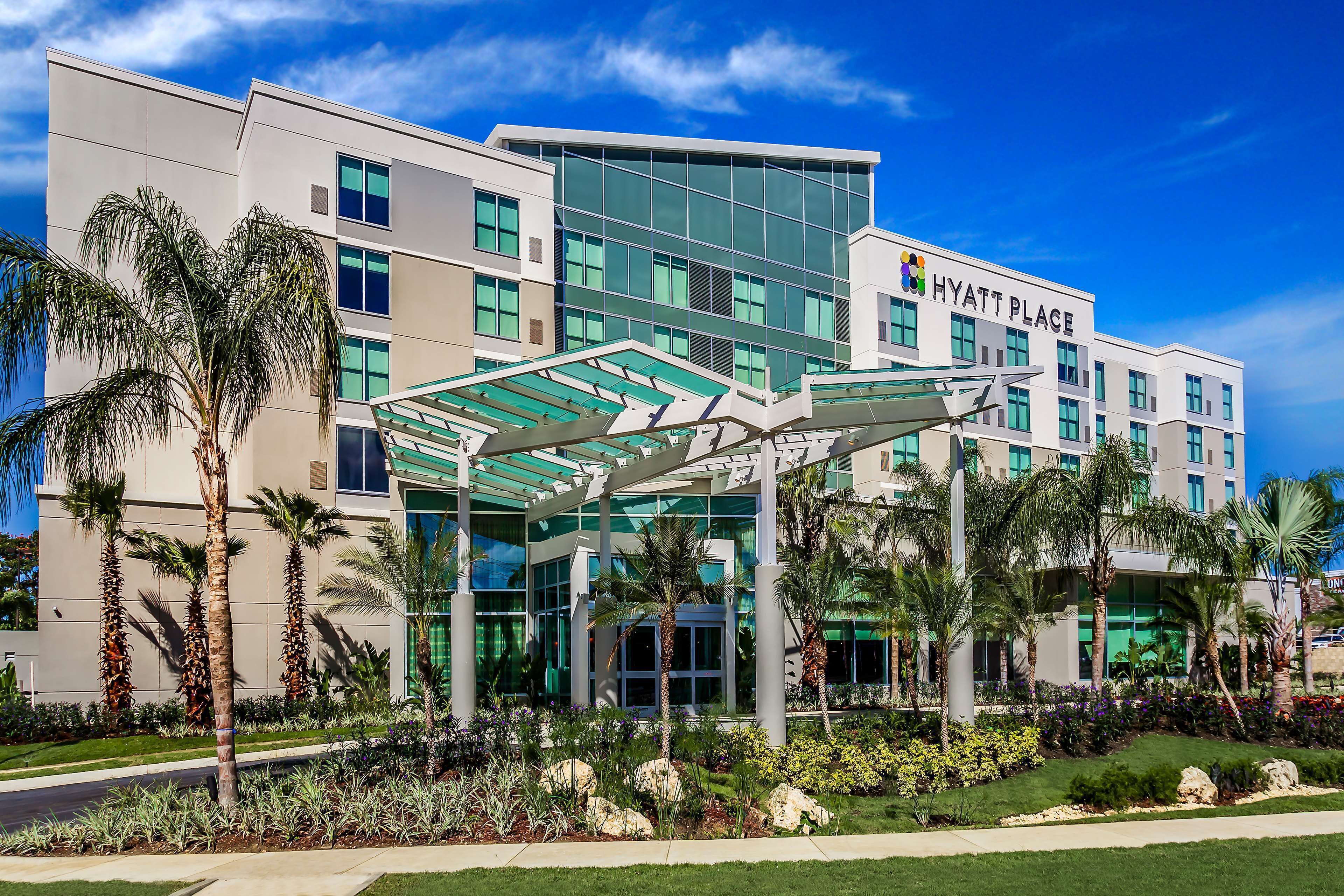 A modern Hyatt Place hotel with a glass entrance, palm trees, and a well-maintained garden.