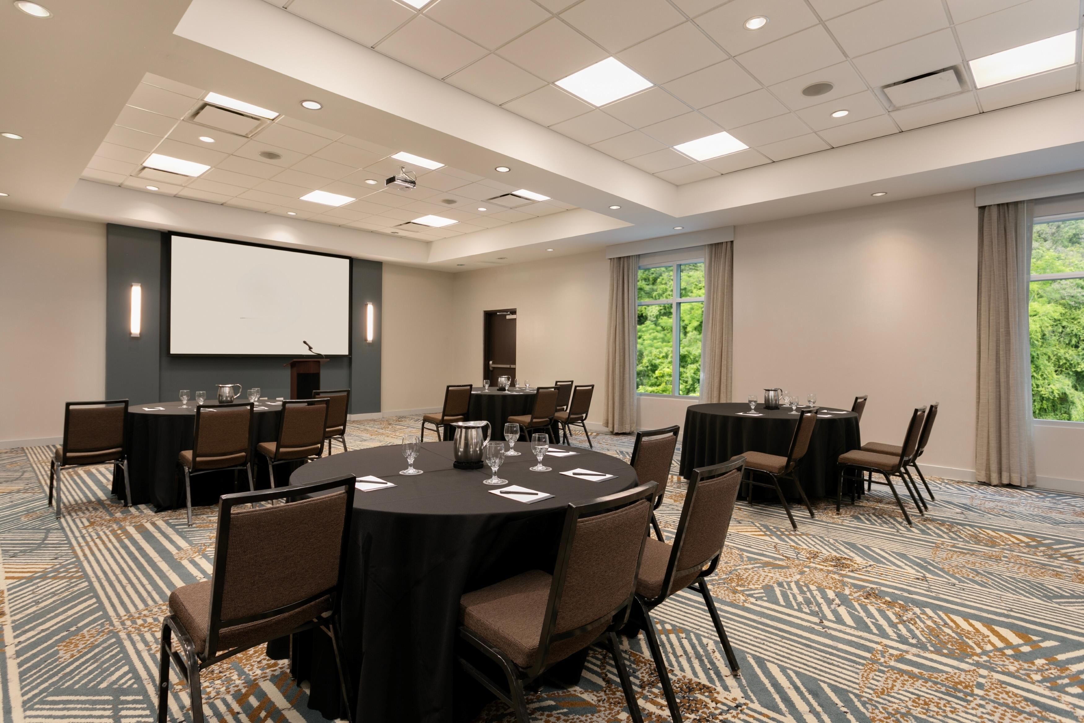 A conference room with multiple tables, chairs, a projector screen, and a large window with a view of greenery.