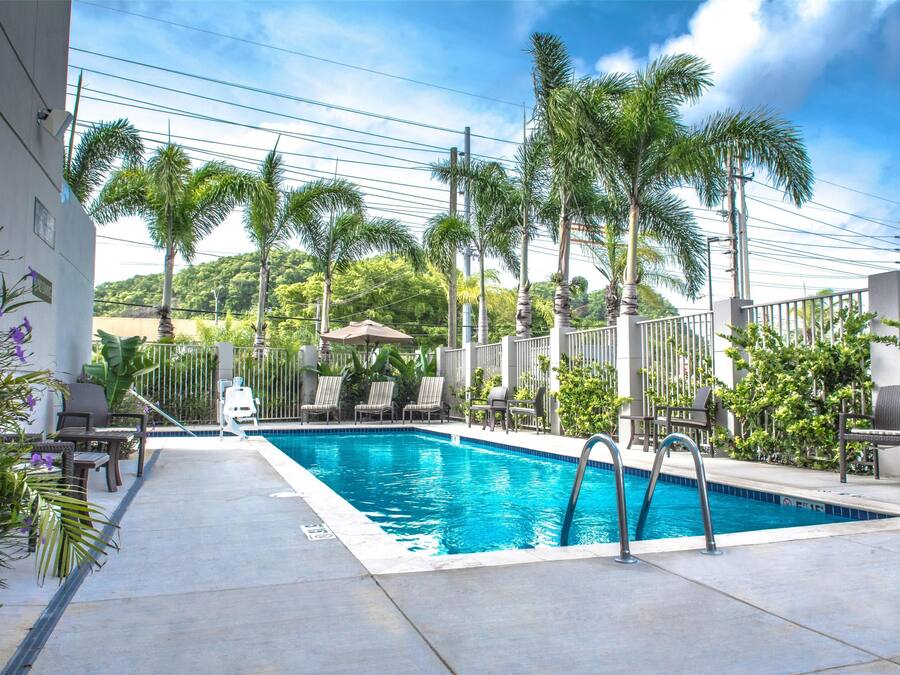 Una piscina rectangular con una escalera metálica, rodeada de sillas de descanso y palmeras, con vista a un paisaje montañoso al fondo.