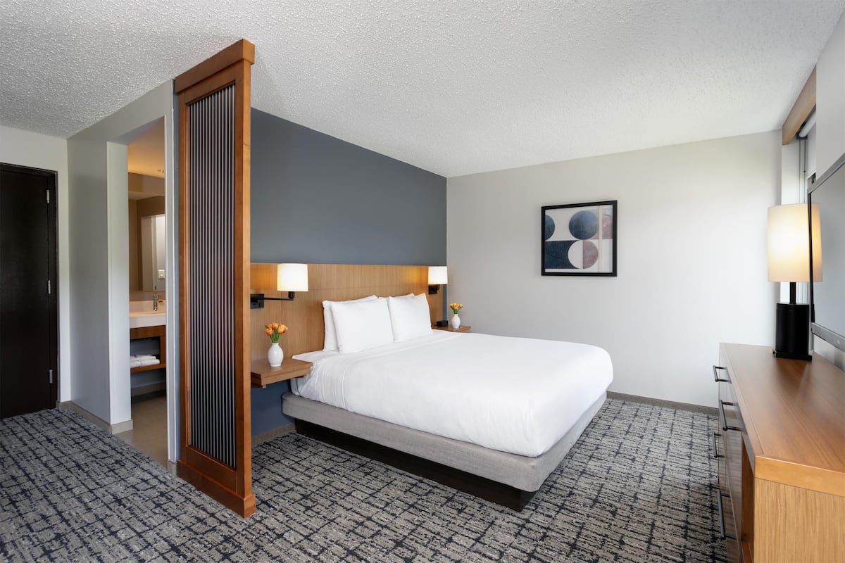 A hotel room with a large bed, a wooden headboard, a desk, a chair, a nightstand, a lamp, a framed picture on the wall, and a bathroom visible through an open door.