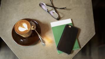 A cup of coffee with a heart design on a saucer, a spoon, a pair of glasses, a green book, and a black smartphone on a table.