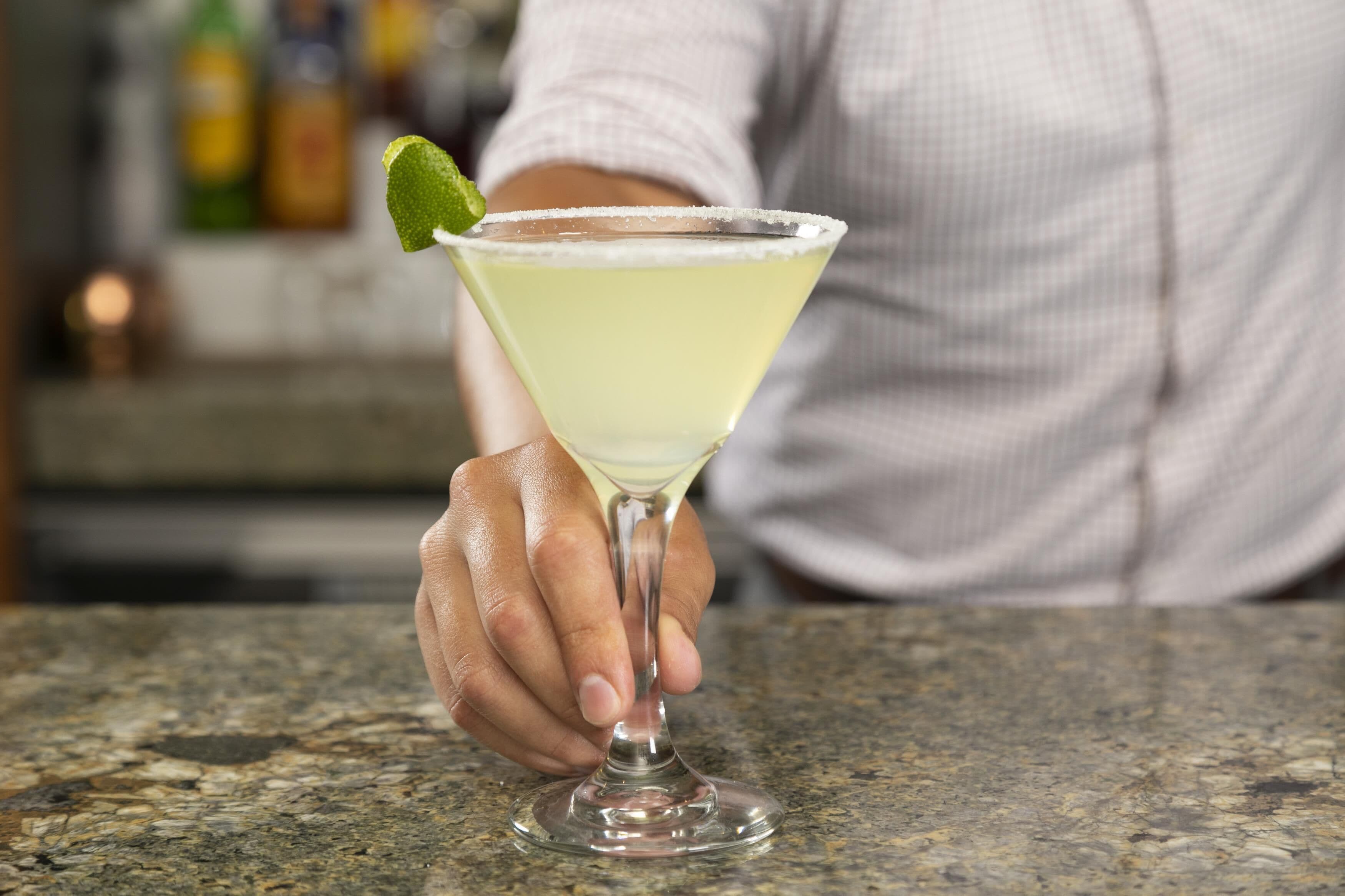 Eine Hand hält ein Martini-Glas mit einer Limettenscheibe auf einer Granitabdeckung.