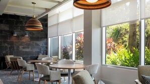 A modern dining area with round tables and chairs, large windows with a view of greenery, and a stone wall. - Hyatt Place Manati (Manati)