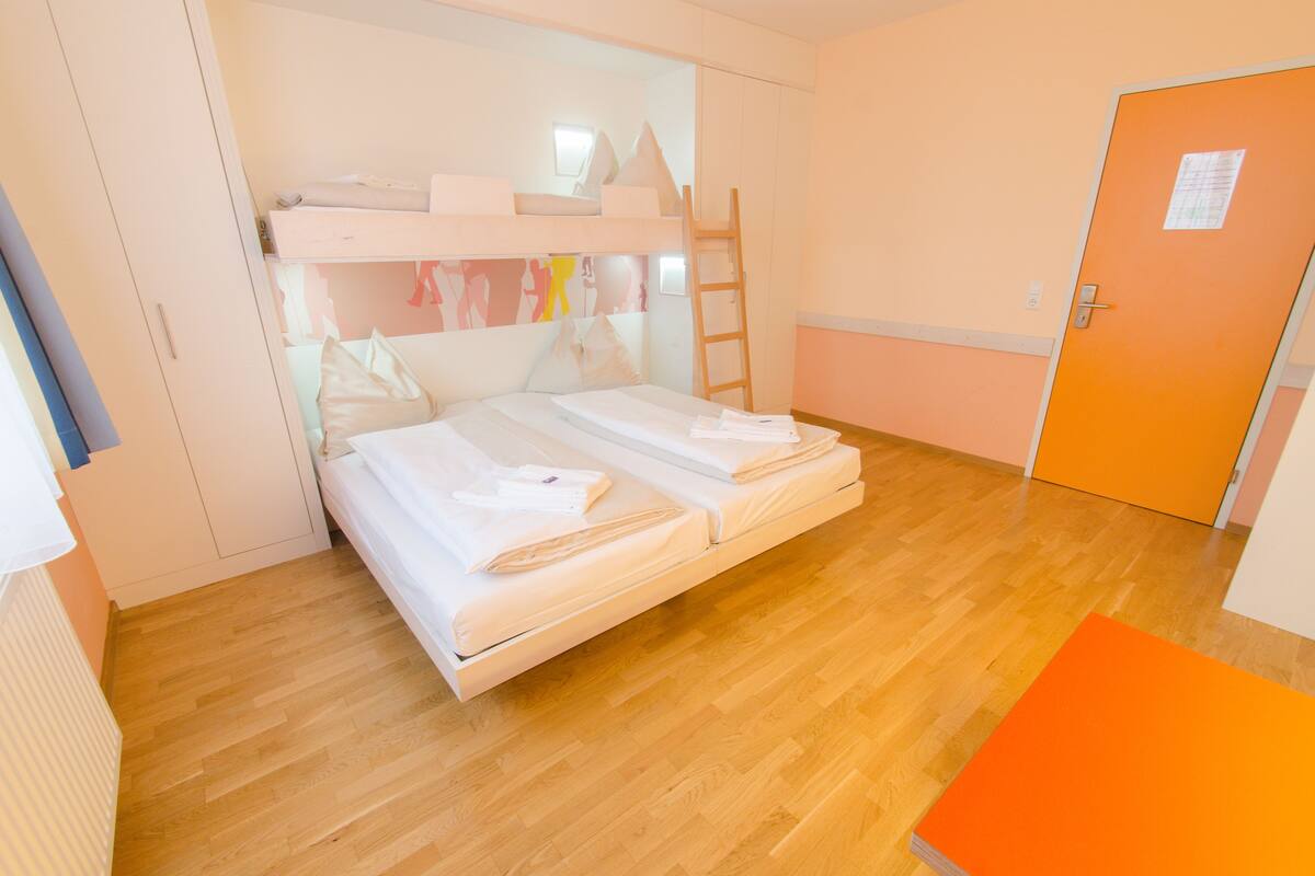 Um quarto com beliche, piso de madeira, escada e porta laranja.