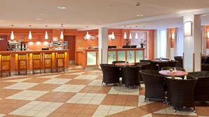 Un vestíbulo de hotel con piso a cuadros, una zona de bar con iluminación anaranjada y mobiliario de mimbre.
