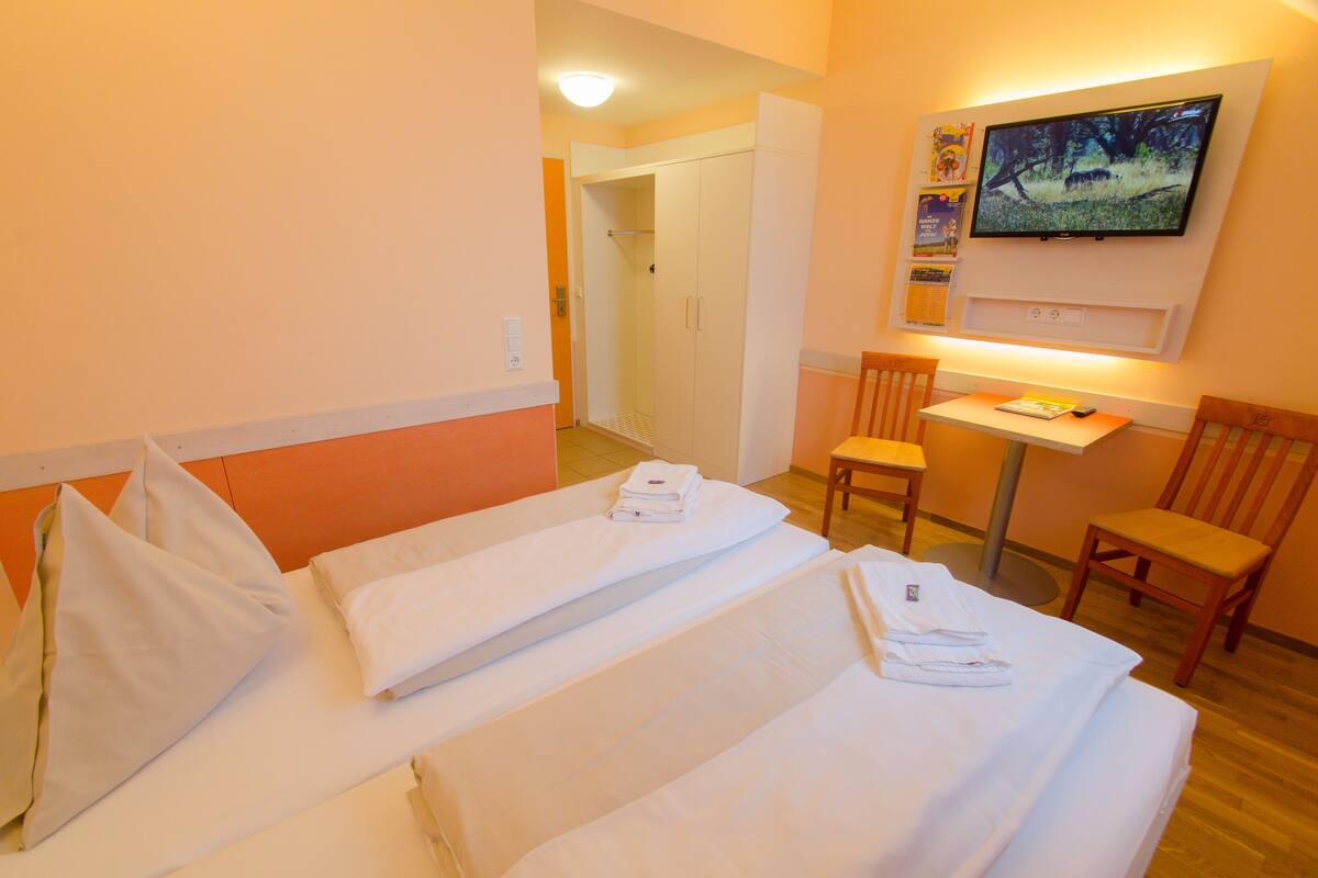 Quarto de hotel com duas camas, uma mesa, uma cadeira, uma TV fixada na parede e uma porta que dá para outro quarto.
