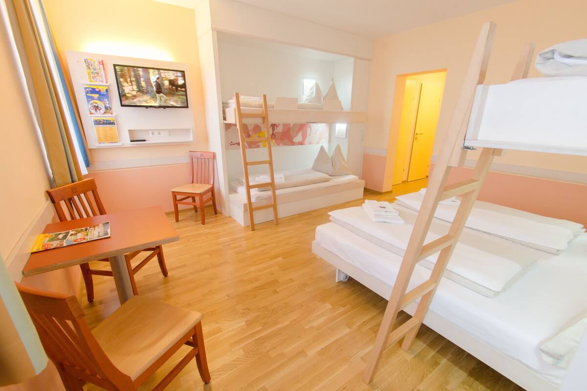Um quarto com beliches, uma mesa de madeira com cadeiras, um pequeno sofá e uma televisão fixada na parede.