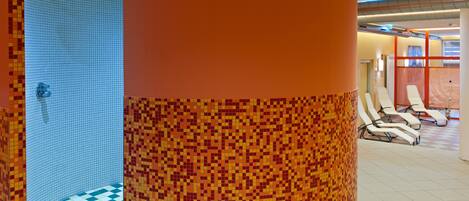 Un baño con zona de ducha con mosaicos, un pilar grande color naranja y una sala de estar con sillas blancas.