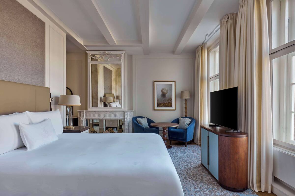 A hotel room with a large bed, a TV, a desk with a lamp, two chairs, and a fireplace.