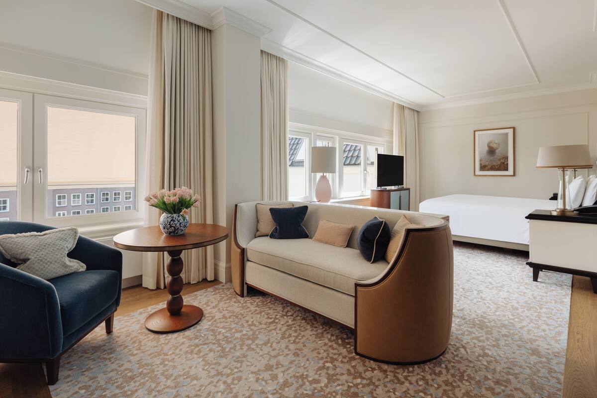 A hotel room with a sofa, armchair, round table, TV, bed, and a painting on the wall.