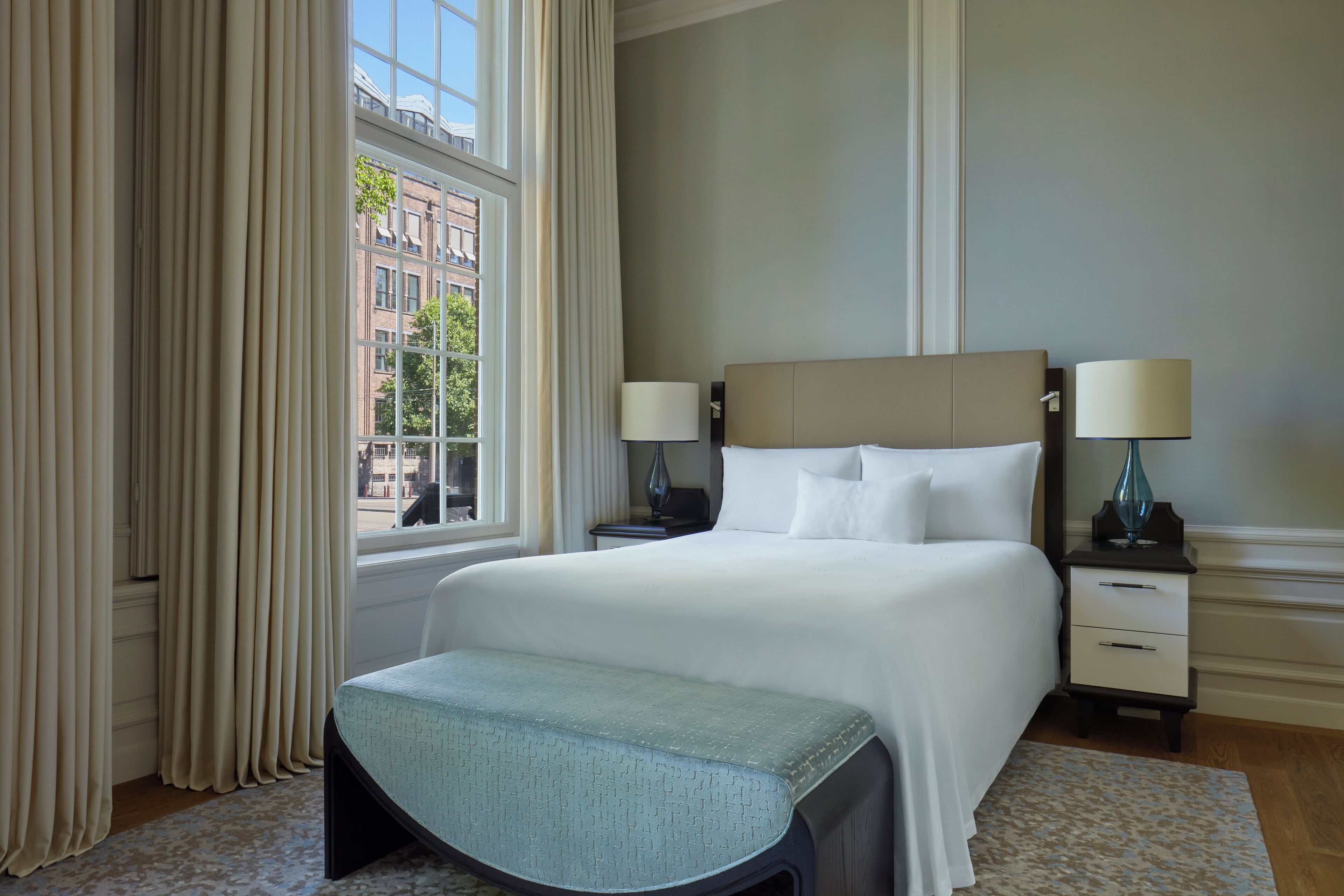 A hotel room with a large bed, two bedside tables with lamps, a bench at the foot of the bed, and a window with curtains.